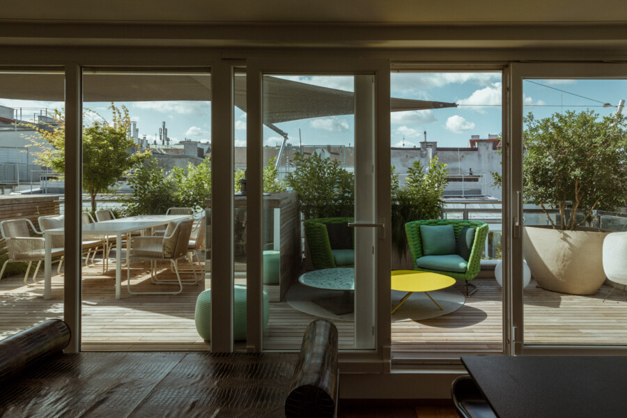 Interior living space with sliding glass doors opening to a sunlit rooftop terrace, featuring wicker seating, a yellow coffee table, and potted shrubs.