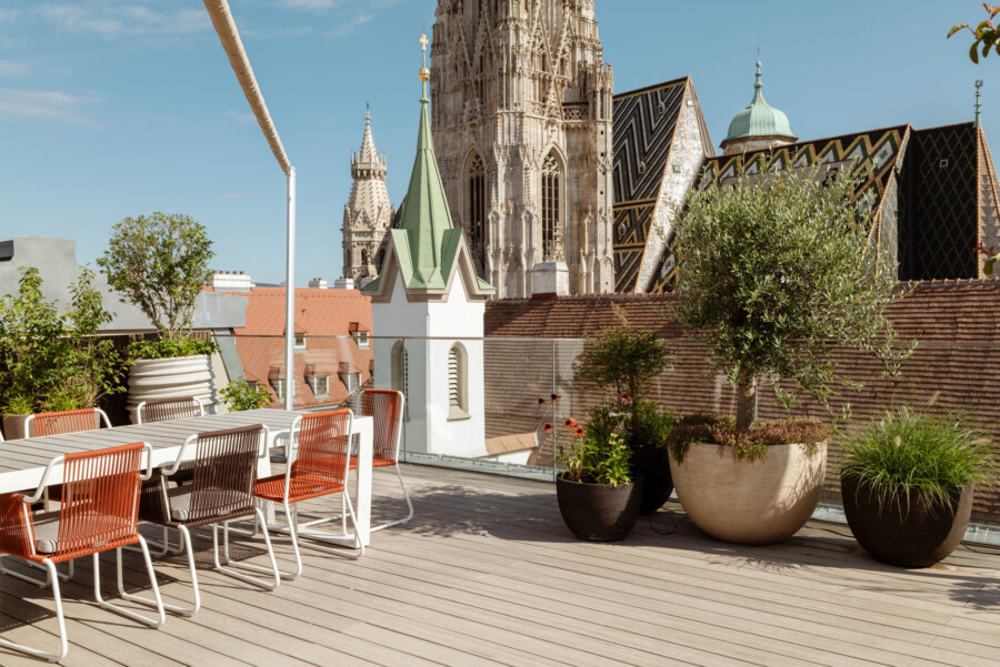 Dachterrasse mit weißen Tischen und orangefarbenen Stühlen, Olea europaea in Töpfen vor gotischer Kathedrale mit buntem Dach.