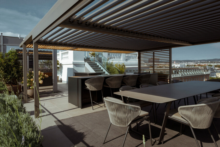 Rooftop patio with a dark slatted pergola, long outdoor dining table and chairs, a bar counter, potted plants, and a distant city skyline.