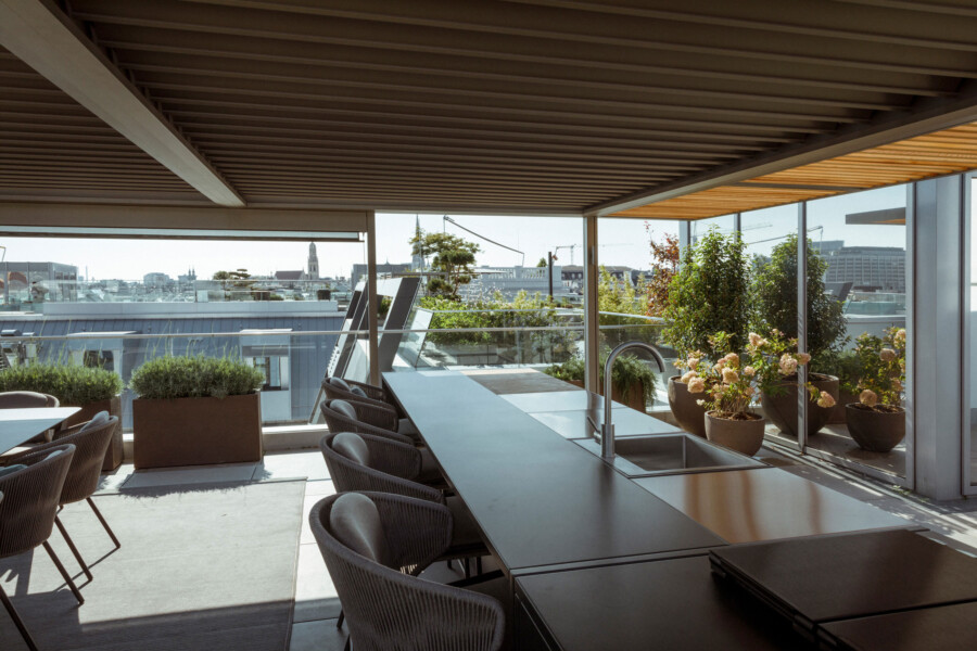 Rooftop terrace with a long counter, sink, and seating, glass railing, potted plants, and a distant city skyline under a slatted wooden ceiling.