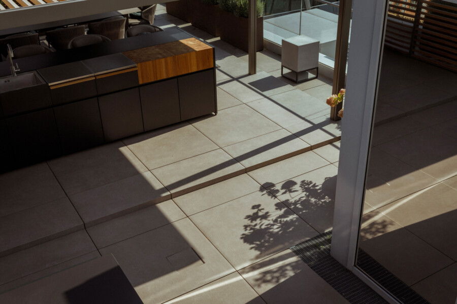 Modern outdoor kitchen with dark cabinets, wooden countertop, and sink; sunlit tiled patio with shadows, a white cube seat, and a potted plant.