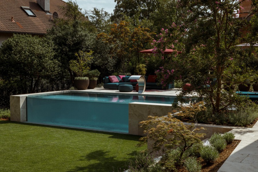 Modern backyard with a rectangular turquoise pool, a teal outdoor sofa with pink cushions on a stone deck, a red umbrella, and lush trees and potted plants.