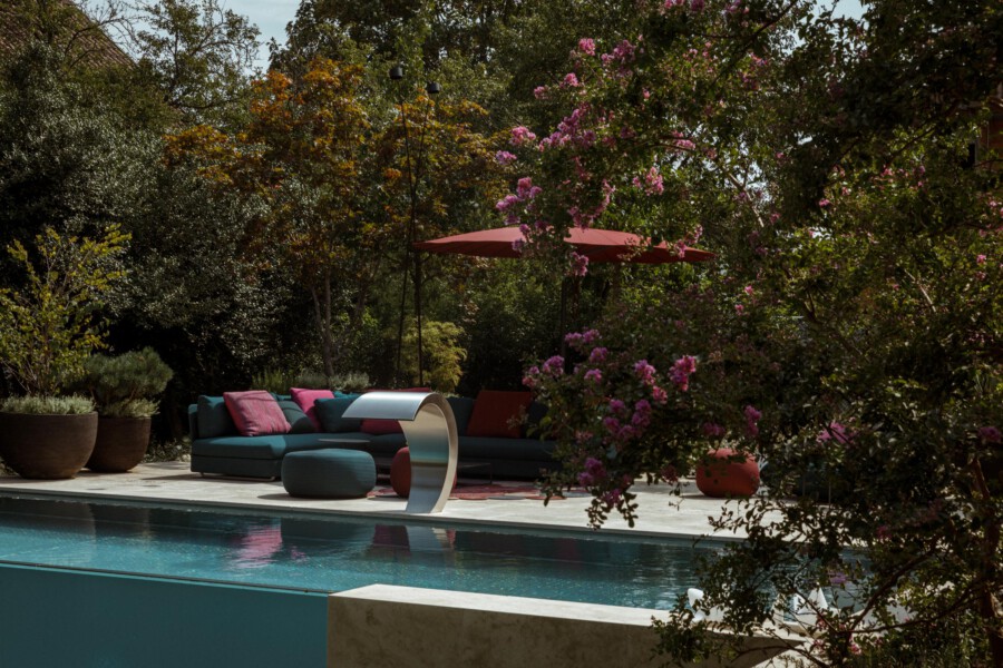 Poolside outdoor lounge with a teal sectional, pink cushions, and a pink umbrella, surrounded by potted plants and flowering shrubs; a curved metal sculpture sits by the pool.