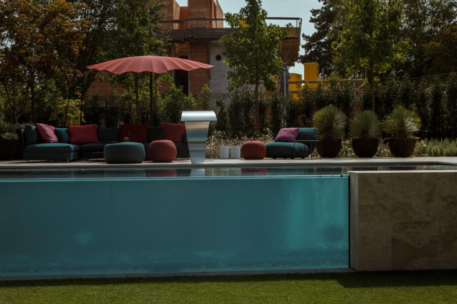 Poolside patio with teal pool, colorful modern lounge seating under a pink umbrella, round poufs, and potted plants; construction behind the garden.