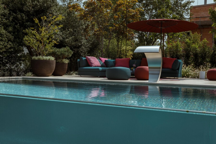 Poolside lounge with a teal sectional, colorful cushions, round poufs, a curved metallic canopy, a red umbrella, and large potted plants among trees.