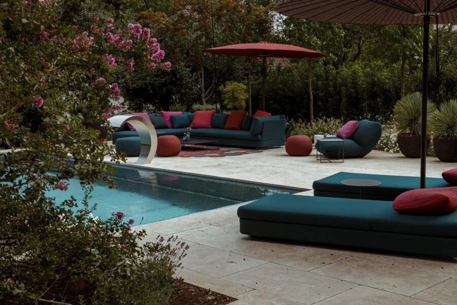 Poolside lounge with teal sectional and chaise, pink/red cushions, red-orange ottomans, curved metal sculpture, large umbrellas, and lush greenery.