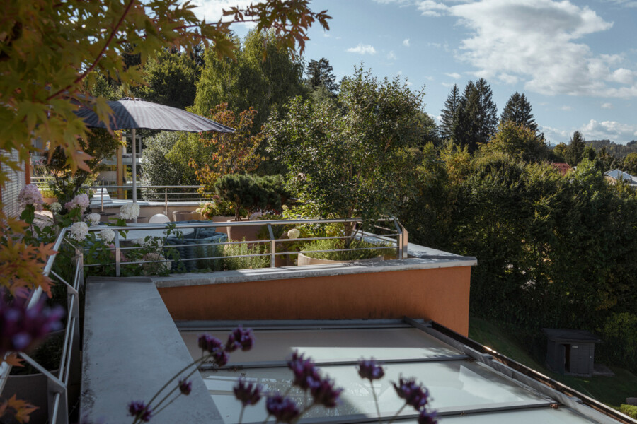 Dachterrasse mit orangefarbener Mauer, Metallgeländer, Sitzgelegenheiten, Sonnenschirm und viele Topfpflanzen; umgeben von Bäumen unter blauem Himmel.