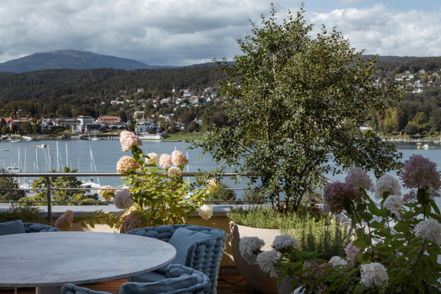 Terrasse mit rundem Marmortisch und blauen Korbsesseln, Hortensien in Pflanzkübeln; Blick über den See mit Booten auf Uferhäuser und dahinter Berge.