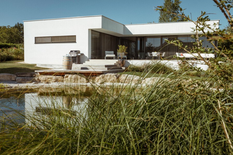 Modern white flat-roof house with large glass doors and horizontal shutters, a deck with seating and a grill, and a pond with tall reeds in the foreground.