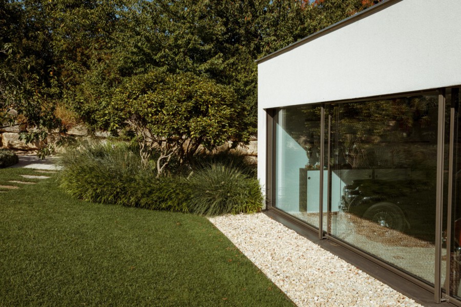 Modern white building with large glass windows, a gravel strip along the base, and a manicured green lawn bordered by shrubs and trees.
