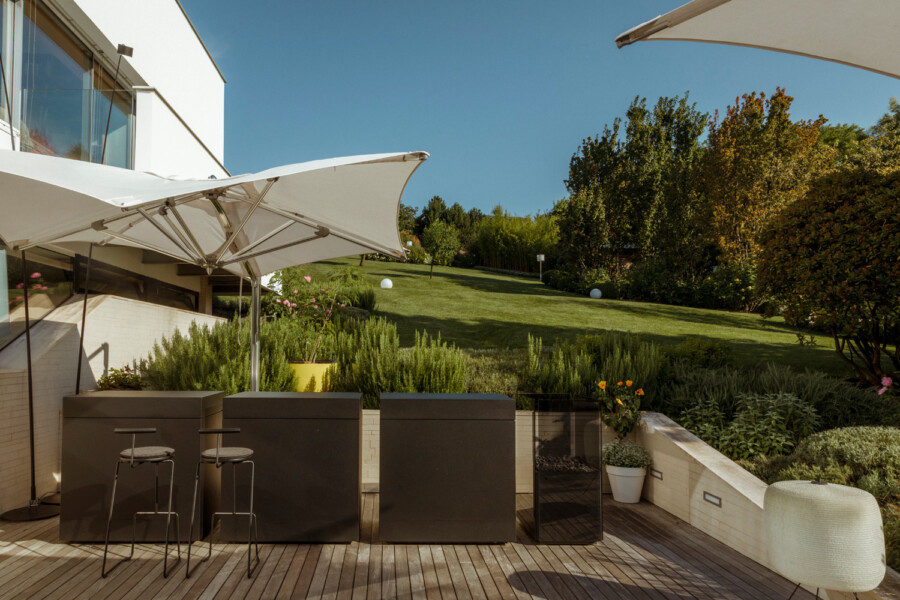 Modern outdoor patio on a wooden deck with white umbrellas, a dark bar counter and stools, potted plants, and a green lawn lined by trees.
