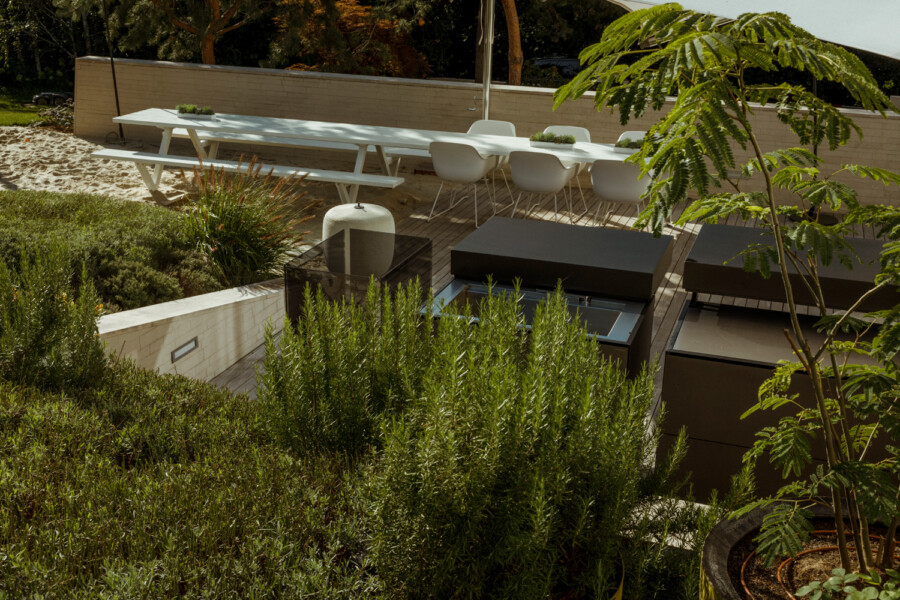 Outdoor modern patio on a wooden deck with a long white dining table, white chairs, brown lounge blocks, and surrounding greenery.