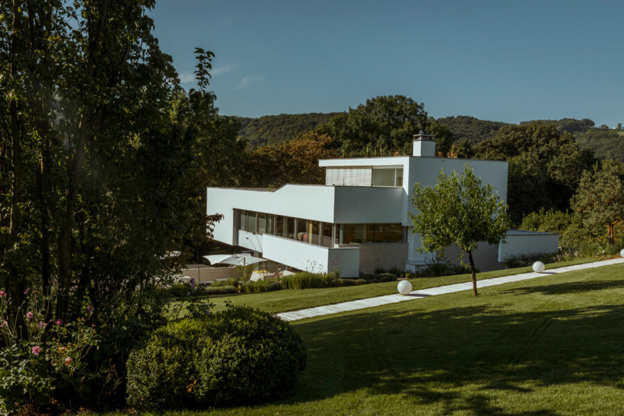 Modern white angular house with glass walls on a green lawn, along a paved path with orb lights, trees and distant hills under a blue sky.