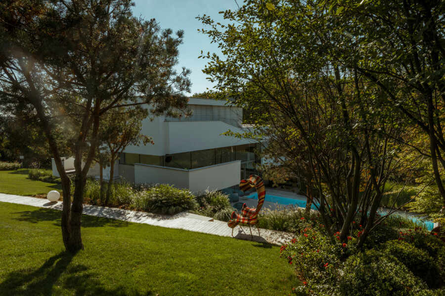 Modern white, flat-roofed building surrounded by trees, with a turquoise pool and a colorful striped sculpture along a paved path in a sunny garden.