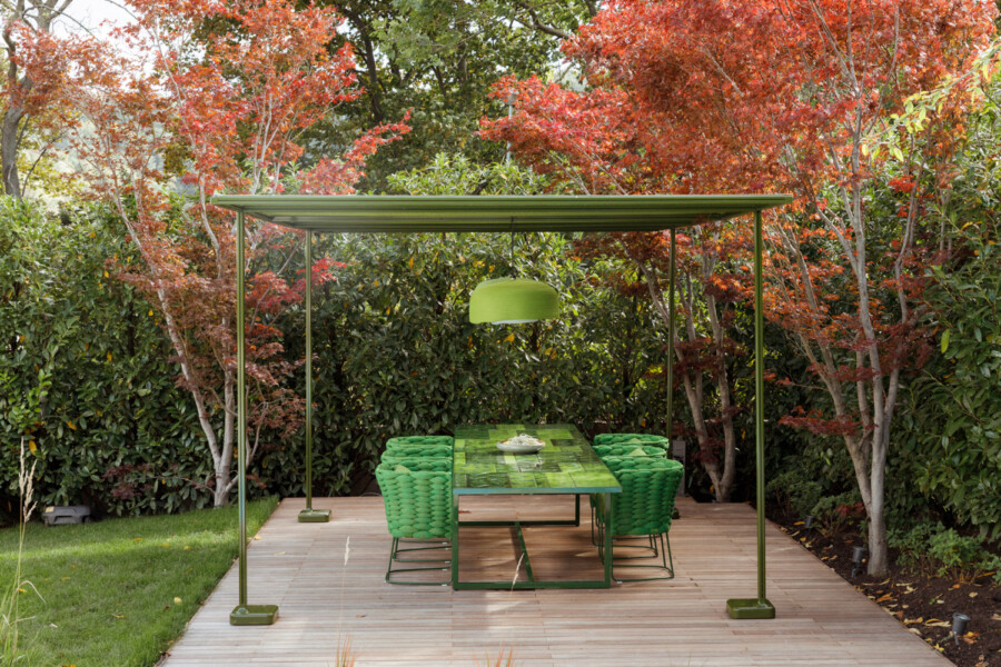 Outdoor dining area under a green metal canopy with a mosaic green table, woven green chairs, and a hanging green lamp, on a wooden deck amid red autumn trees.