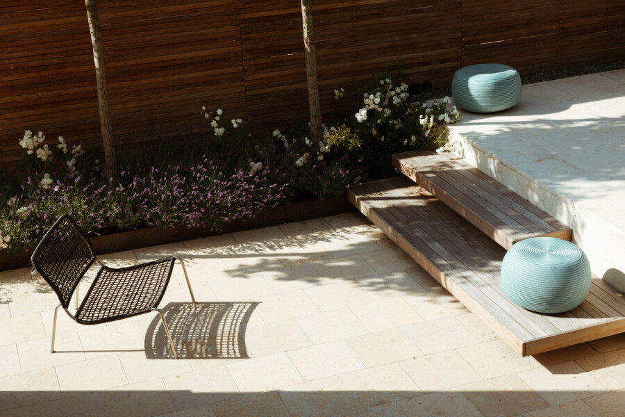 Sunny outdoor patio with pale tiled floor, a black wire chair, wooden steps, two turquoise poufs, and a flower bed along a wooden fence.