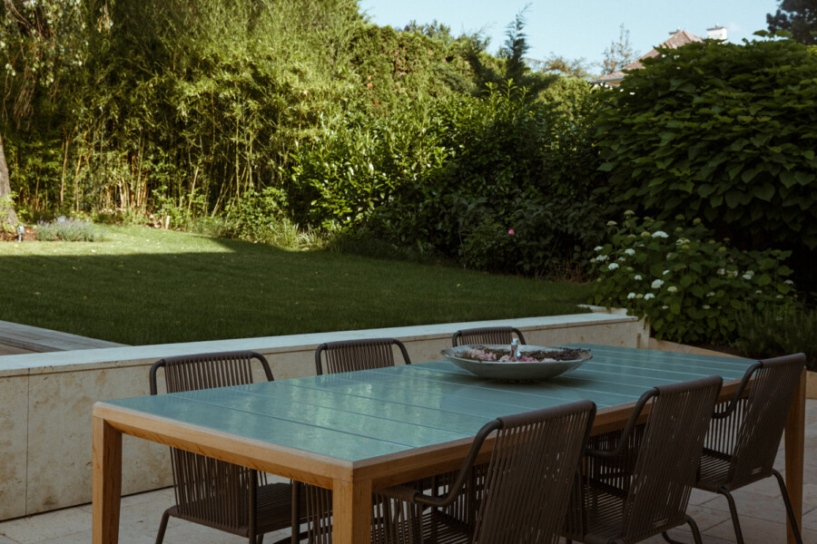 Outdoor dining area with a glass-topped wooden table and brown wicker chairs on a stone patio, a large bowl centerpiece, and a lush green lawn with hedges.