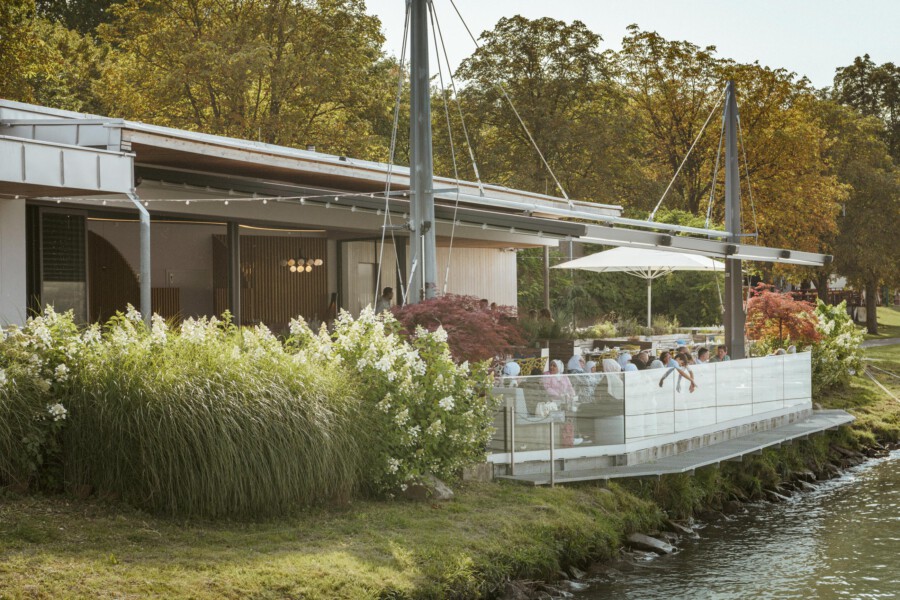 Flussufer-Restaurant mit Glasbalkon; Gäste speisen draußen, davor Grasstauden mit weißen Blüten, Herbstbäume im Hintergrund.