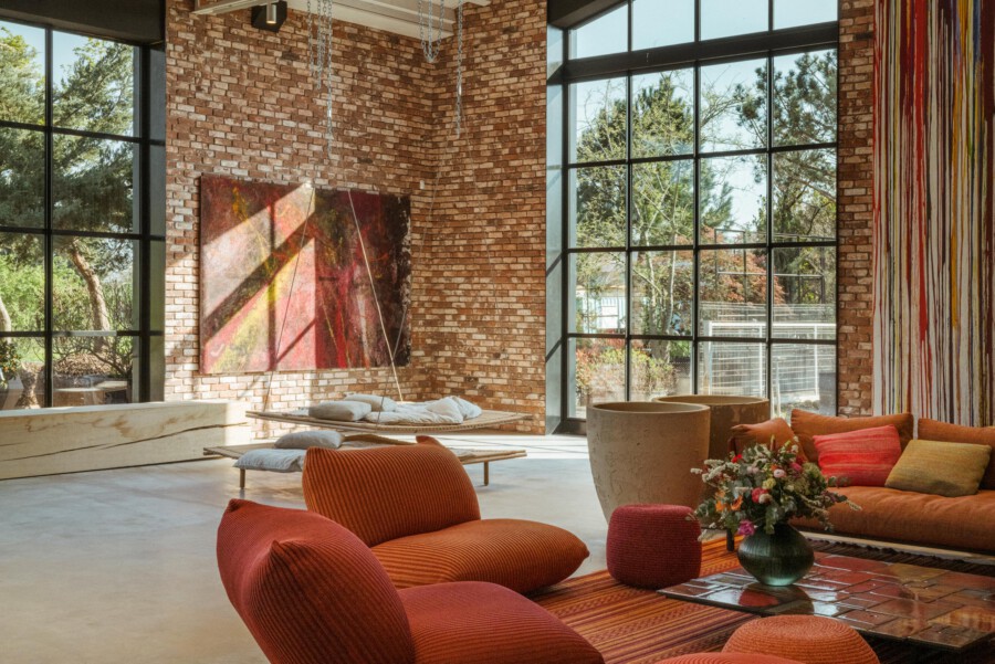 Loft-Wohnzimmer mit Backsteinwand und großen Fenstern; orange Sitzmöbel, Blumenarrangement in Vase auf Couchtisch, abstrakte Wandkunst.