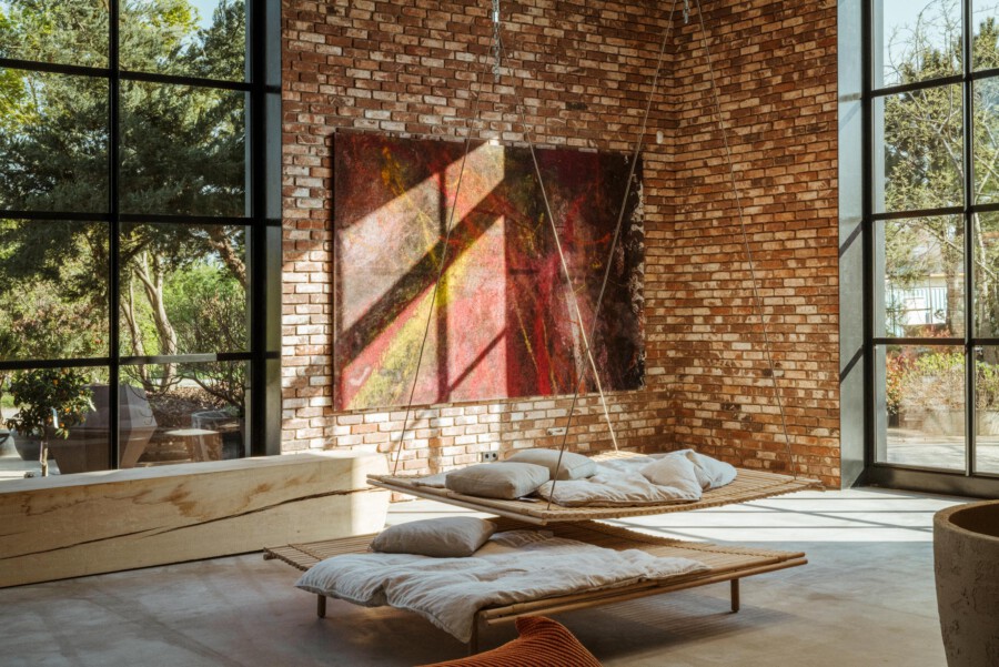 Industrielles Wohnzimmer mit Ziegelwänden und großen Fenstern; Holzliege mit Kissen; abstraktes Gemälde; Blick auf Gartenpflanzen außerhalb.