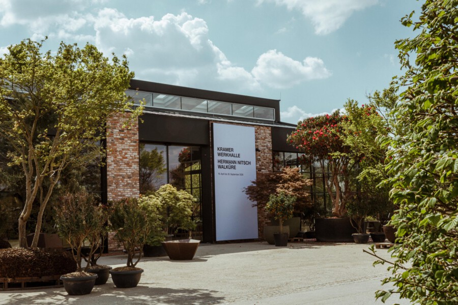 Außenansicht eines Bruchstein-Gebäudes mit großen Fenstern; Topfpflanzen und Bäume im Vorgarten; Schild: Kramer Werkhalle Hermann Nitsch Walküre.
