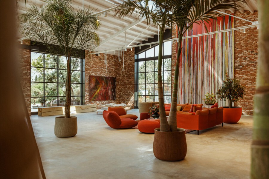 Innenlounge mit orangefarbenen Sitzmöbeln; Dypsis lutescens-Palmen in Terrakotta-Töpfen vor Ziegelwand und großen Fenstern.