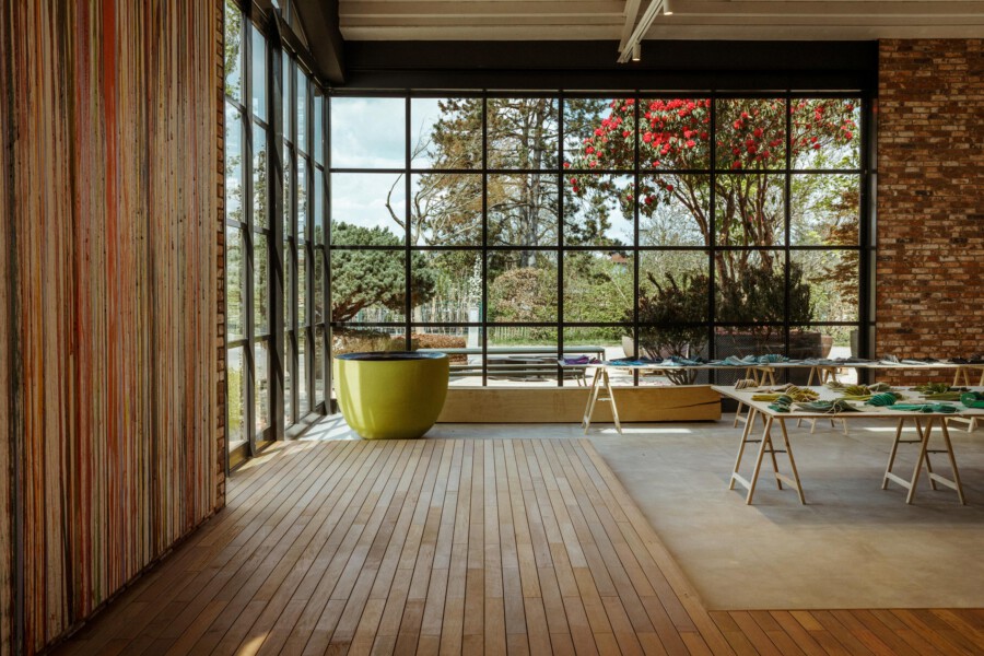 Innenraum mit großem Glasfensterblick; Rhododendron spp. mit roten Blüten im Garten; Holzfußboden, Ziegelwand, moderne Möbel.