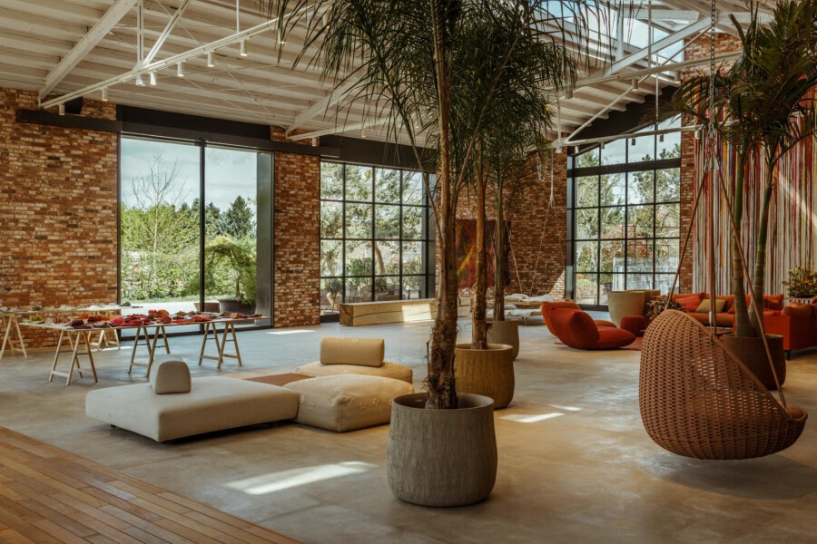 Offener Innenraum mit Backsteinwänden und großen Fenstern; Dypsis lutescens-Palmen in Töpfen; gemütliche Lounge mit orangefarbenen Sesseln.