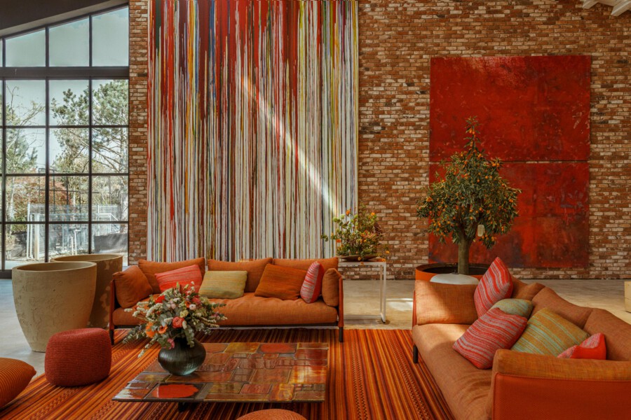 Modernes Wohnzimmer mit orangefarbenen Sofas; Orangenbaum Citrus sinensis im Topf vor roter Wand und Ziegelwand, große Glasfront.