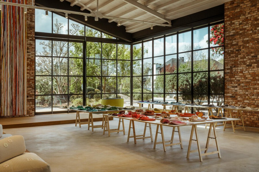 Großer Innenraum mit Glasfront; lange Tische mit farbigem Geschirr; gelbe Topfpflanze am Fenster, Ziegelwände.