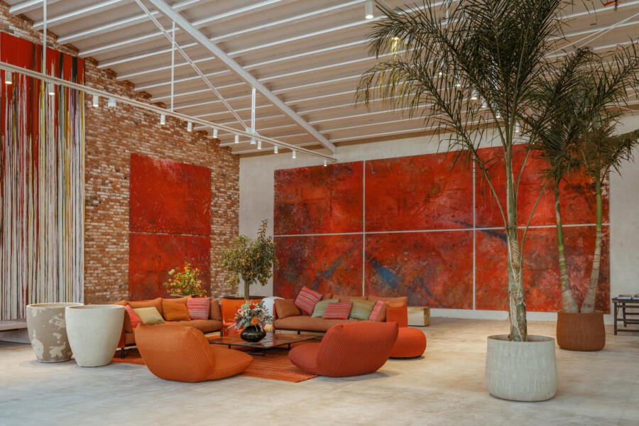 Innenraum einer Kunstgalerie mit orangefarbenen Sitzmöbeln, roten abstrakten Wandbildern, Backsteinwänden und Dypsis lutescens-Palmen.