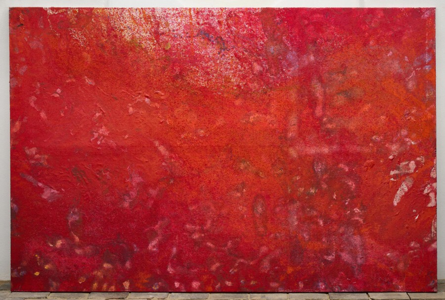 Abstraktes rotes Gemälde mit texturierter Oberfläche, lehnt an grauer Wand auf gepflastertem Boden.