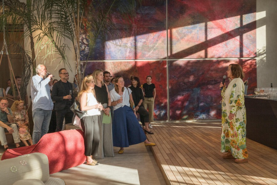 Gruppe von Anwesenden hört einer Person mit Mikrofon zu; rechts spricht diese Person, links Palmen (Dypsis lutescens) vor farbiger Wandkunst.