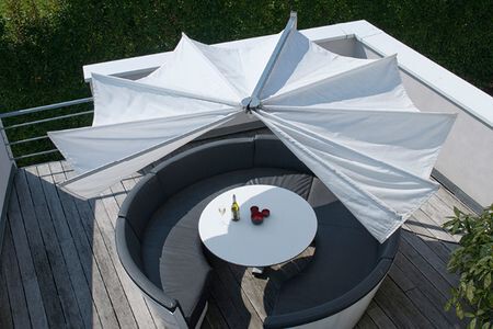 Circular outdoor seating around a white round table on a wooden deck; large white umbrella opened; bottle and red fruit on table; leafy hedge border.