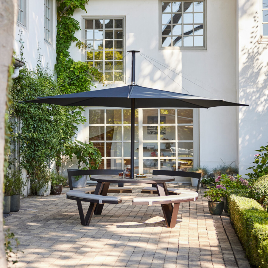 Round wooden table with curved benches under a large dark umbrella in a paved courtyard; white-wall house, Hedera helix ivy, potted plants, and a trimmed hedge.