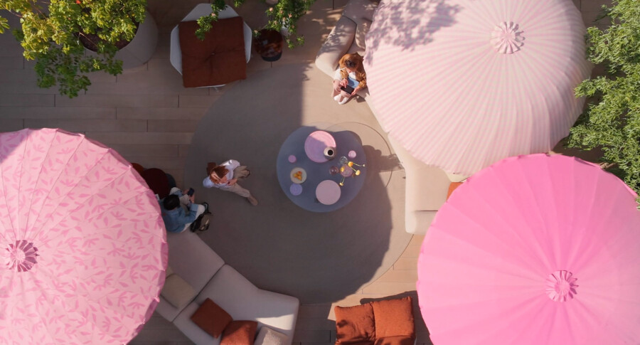 Top-down view of a rooftop outdoor lounge with pink parasols, beige sofas, and potted greenery; people gather around a round coffee table.