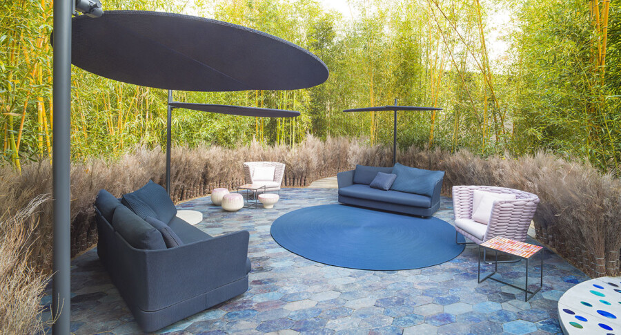 Outdoor lounge area with gray and blue sofas around a circular blue rug; shade sails overhead; tall bamboo (Bambusa sp.) in the background.