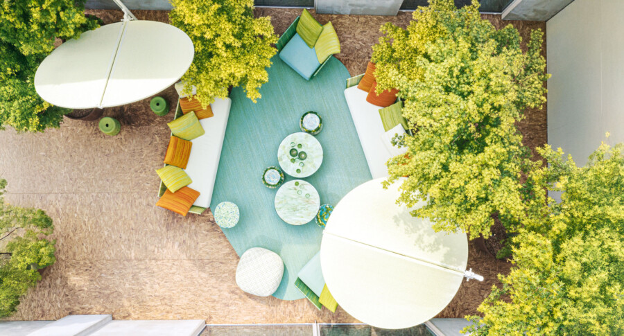 Overhead view of circular outdoor lounge with white sofas on a teal rug, multicolor cushions, and golden-leaved shrubs surrounding.