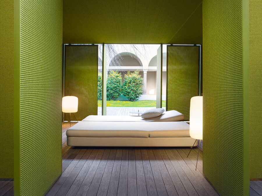 Grünes Schlafzimmer mit weißem Bett, Holzboden und zwei Stehlampen; Blick durch Glasfront auf Garten mit Sträuchern.