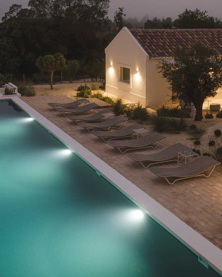 Poolbereich mit Liegen, kleines Haus mit Außenbeleuchtung; Garten mit Trockenpflanzen wie Yucca sp. und Gräsern bei Dämmerung.