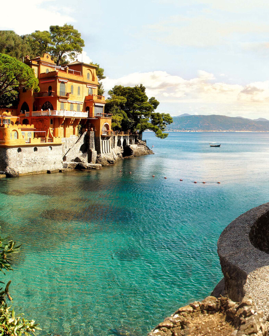 Orangefarbene Villa auf Felsen ragt ins Mittelmeer; von Pinus pinea umgeben; türkisblaues Wasser, Boot im Hintergrund.