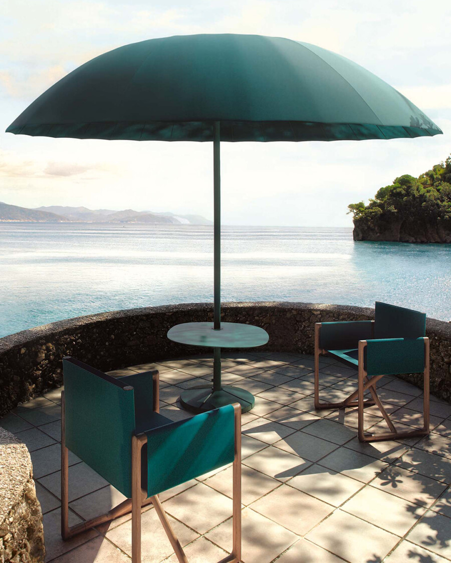 Sitzplatz am Meer unter grünem Sonnenschirm, runder Beistelltisch und drei Teakholz-Sessel, Blick auf Küste und Insel.