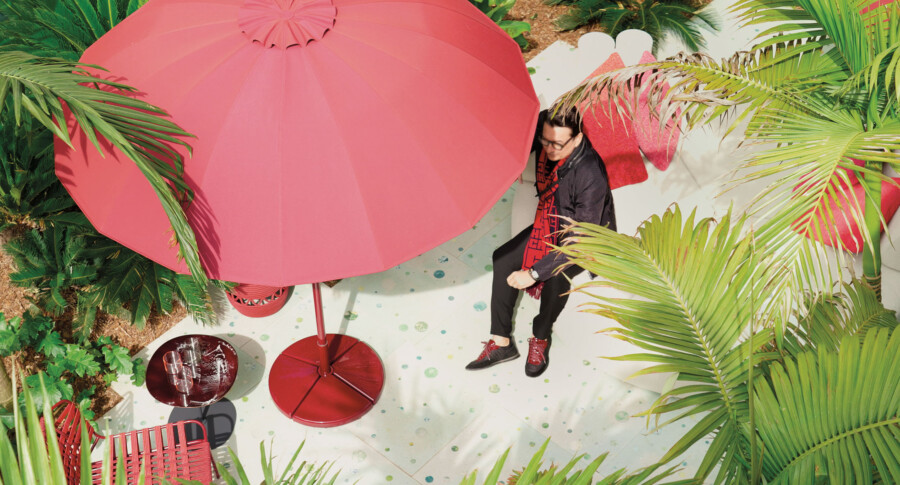 Person seated under a large red umbrella on a tropical patio, surrounded by Arecaceae palms and red outdoor furniture.
