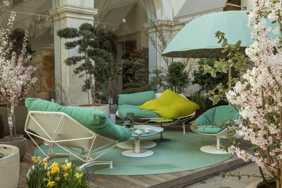 Außenterrasse mit weißen Loungesesseln, grünen Kissen, runden Tischen; türkisfarbener Sonnenschirm; Buxus sempervirens-Topiaria und Narcissus spp.