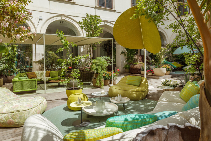 Innenhof-Gartenlounge mit gelben Sitzsäcken und runden Tischen; zahlreiche Topfpflanzen prägen den Freiluftbereich.