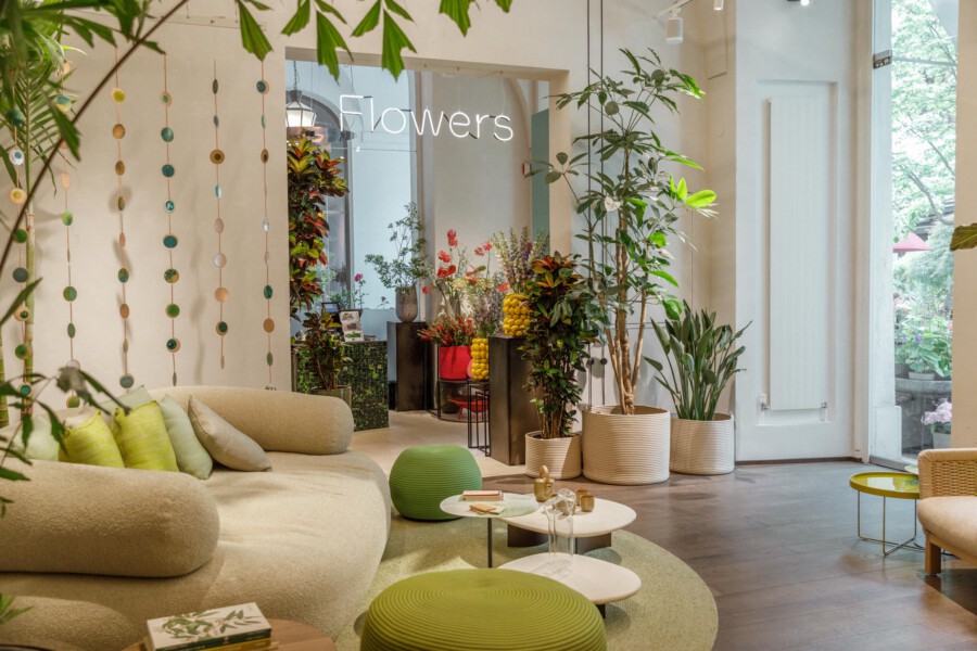 Moderner Innenraum mit Zimmerpflanzen, u. a. Ficus elastica und Dracaena fragrans; Neon-Schriftzug 'Flowers', gemütliche Sitzecke.