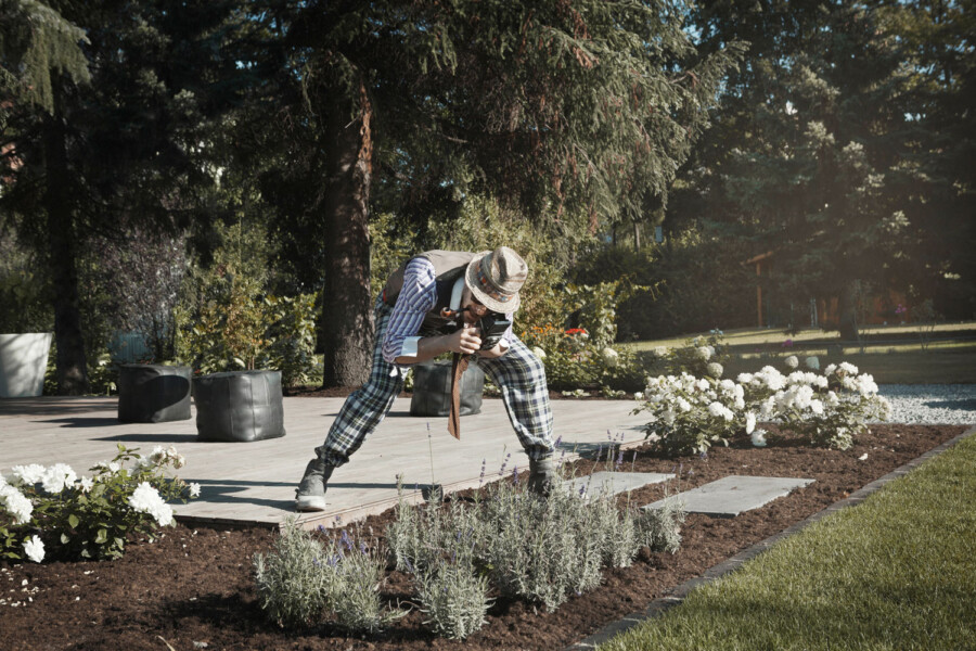 Person mit Strohhut und kariertem Outfit kniet im Garten und fotografiert Blumen mit einer Kamera; im Beet blühen weiße Rosen und Lavendel.