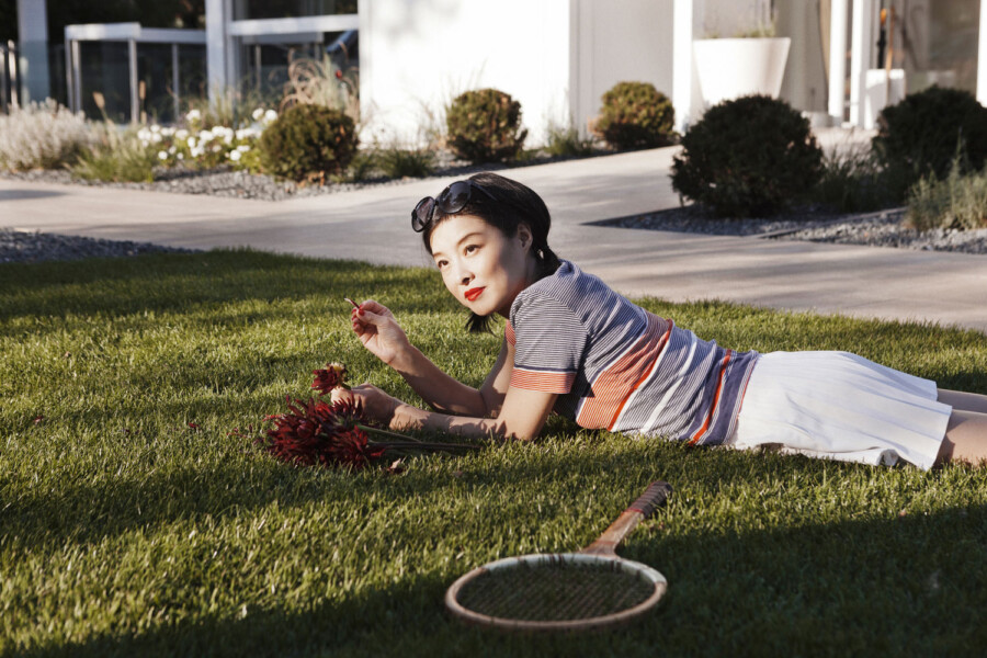 Eine Person liegt auf der Wiese, schaut in die Kamera, hält rote Blumen; neben einem Tennisschläger, trägt gestreiftes Oberteil und weiße Hose.