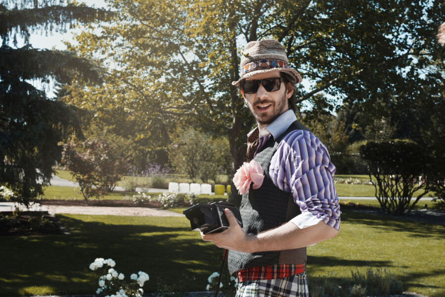 Person im Park trägt Strohhut, Sonnenbrille, eine lila gestreifte Bluse und Weste, rosa Blume am Revers; hält eine Kamera und lächelt.