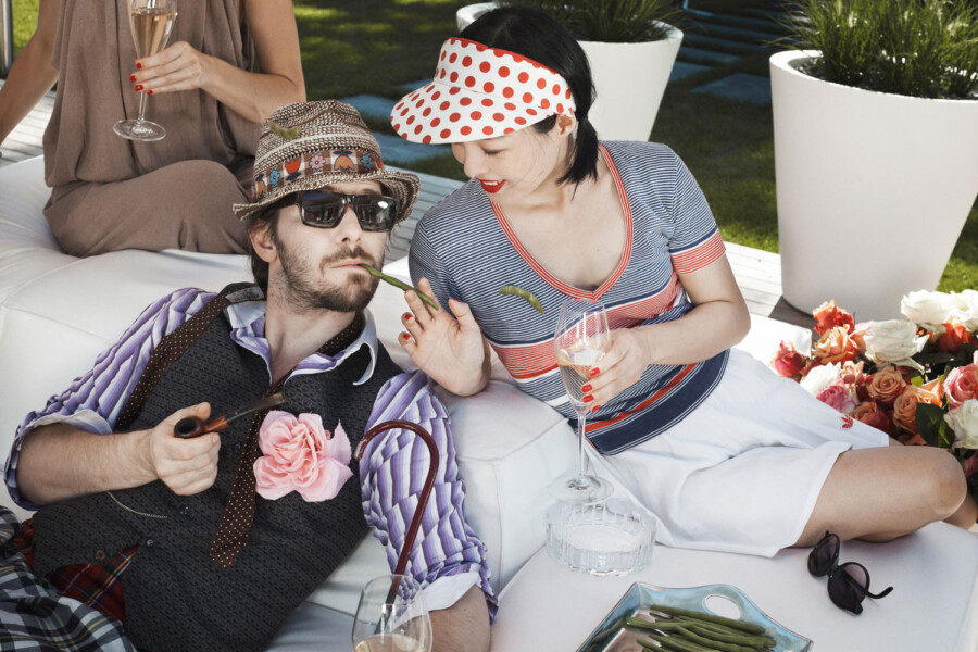 Zwei Personen entspannen sich im Garten auf einer weißen Lounge; eine reicht dem anderen eine grüne Bohne, beide tragen Retro-Outfits.